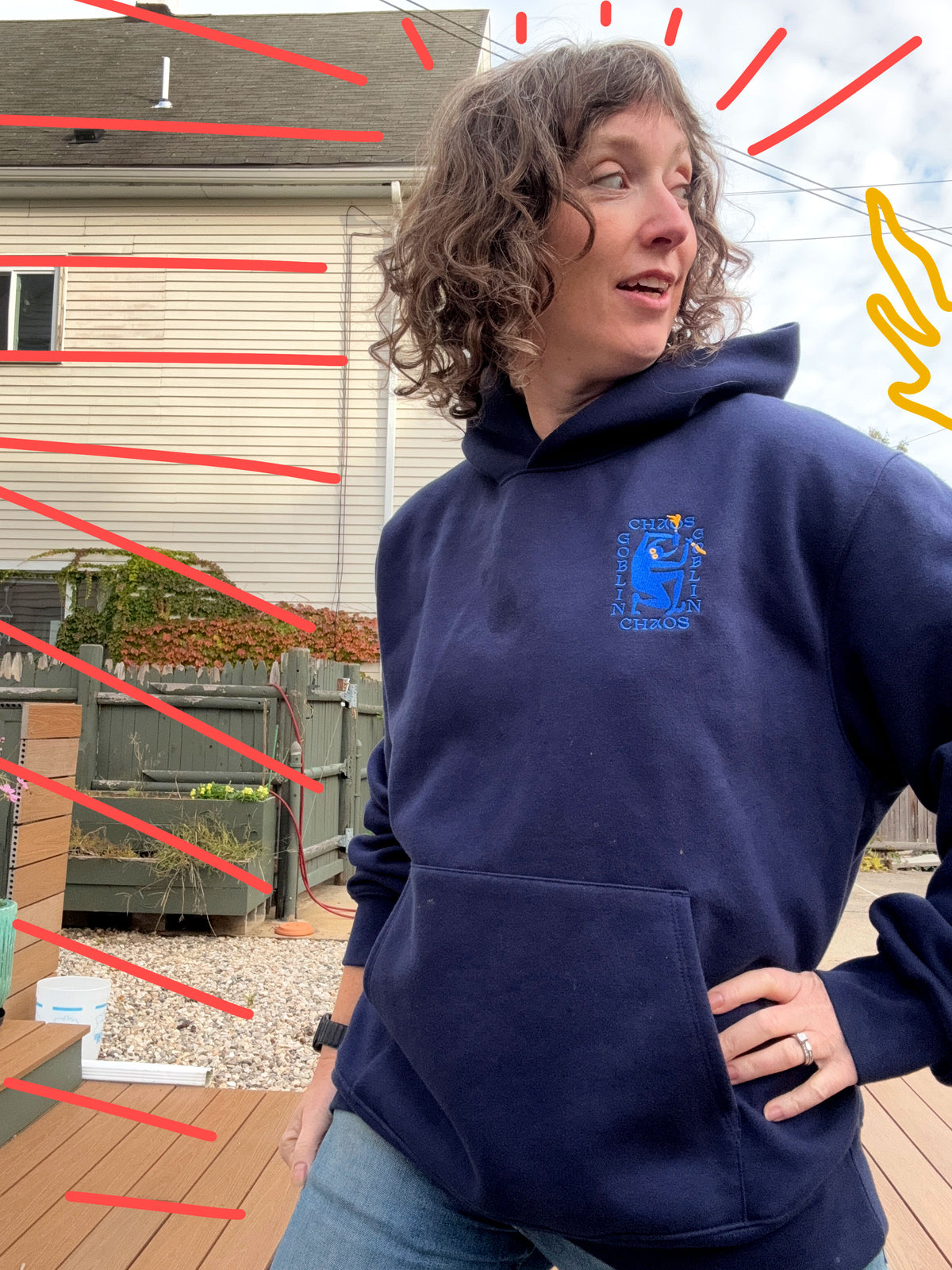 A woman wearing a navy blue hoodie with a blue and yellow 'Chaos Goblin' logo on the left chest in a medieval linocut style.