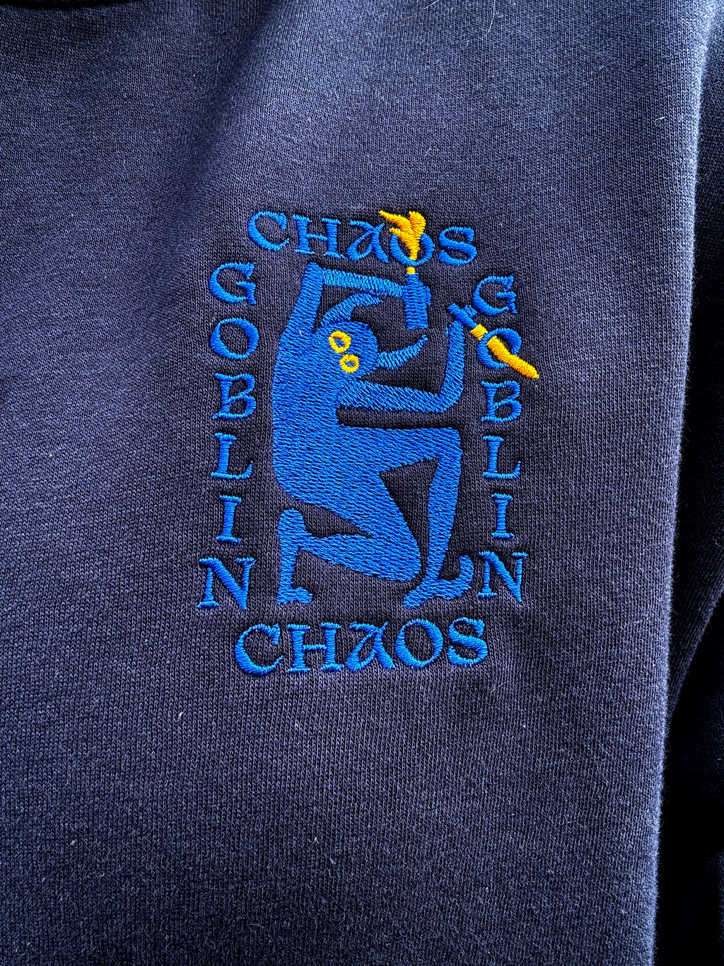 Close-up of a navy blue sweatshirt featuring a blue and yellow embroidered graphic with the words 'Chaos Goblin' and a stylized figure.