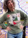 A woman wearing an olive green crewneck sweatshirt with a colorful patchwork quilt design on the front, posing outdoors with her hands on her hips. The sweatshirt features a patchwork of various fabrics including scottie dogs, houndstooth, and several maps.